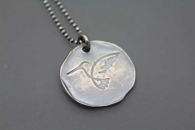 Op de afbeelding: Een zilveren ketting met een ronde hanger met een kolibrie-ontwerp. De hanger heeft een getextureerde, verweerde afwerking.