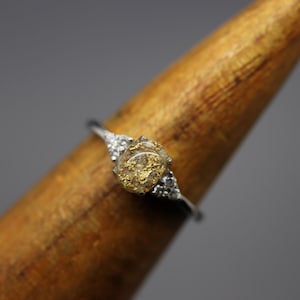 May include: A silver ring with a clear stone set in the center. The stone is filled with small pieces of gold leaf. The ring is set on a wooden display stand.