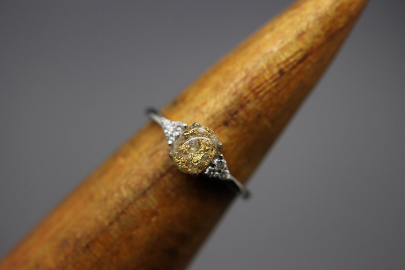May include: A silver ring with a clear stone set in the center. The stone is filled with small pieces of gold leaf. The ring is set on a wooden display stand.