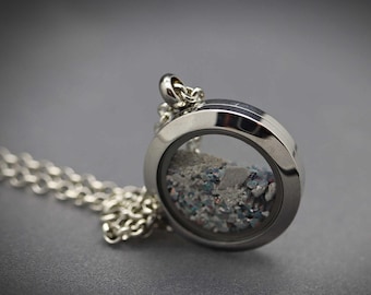 Peut inclure: Un pendentif médaillon circulaire en métal argenté suspendu à une chaîne à maillons assortie. Le médaillon présente une face en verre transparent révélant une collection de petits fragments de pierre concassée gris, bleu et irisé.