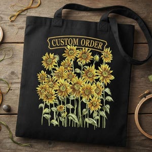 May include: A black tote bag featuring a central graphic of yellow sunflowers with green stems and leaves. Above the floral arrangement, a gold-coloured banner contains the text "CUSTOM ORDER" in a bold, capitalised, serif font style.