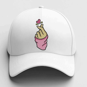 Può includere: Cappellino da baseball bianco con un disegno ricamato di una mano che forma un cuore. La mano ha una manica rosa e il cuore è rosa con dettagli gialli.