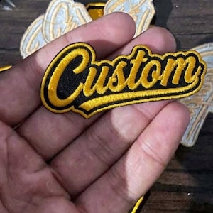 Puede incluir: Un parche bordado que presenta la palabra Custom en una tipografía cursiva y llamativa. Las letras están realizadas con hilo amarillo brillante y un borde negro grueso, creando un diseño de alto contraste para aplicar sobre telas.