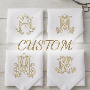 May include: Four white square napkins arranged in a grid, each featuring a unique gold embroidered monogram. The word CUSTOM is centered in gold text. The napkins are folded into points, with a spool of gold thread and scissors visible above.