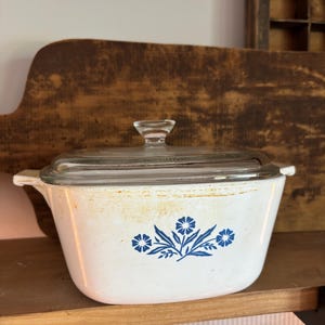 May include: White casserole dish with a clear glass lid. The dish features a blue floral design on the front. The dish is sitting on a wooden shelf.