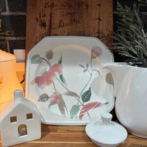 May include: A white ceramic house-shaped candle holder with square windows and a lit candle inside. A floral patterned plate, a white pitcher, and a wooden cutting board with handwritten text are also visible.