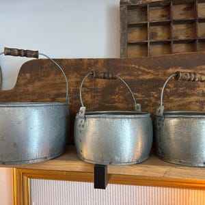May include: Three galvanized metal buckets with wooden handles and wire bails. The buckets are displayed on a wooden shelf against a rustic wooden background. The buckets are of varying sizes, with a weathered appearance.