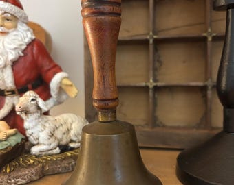 May include: A vintage brass bell with a wooden handle. The bell has a flared base and a dark brown handle. The handle is made of turned wood. The bell is likely used for decorative purposes or as a collectible item.