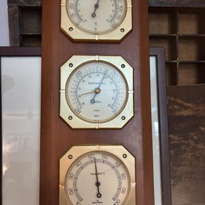 May include: A wooden wall-mounted weather station with three brass-framed gauges. The top gauge is a thermometer, the middle a barometer, and the bottom a humidity gauge. The thermometer displays in Celsius.