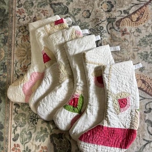 May include: A set of five quilted Christmas stockings in off-white fabric, each featuring unique floral-inspired appliqué patterns in shades of pink, red, green, and beige. Each stocking includes a small white fabric loop for hanging.