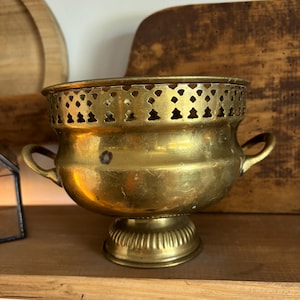 May include: A vintage brass bowl with two small handles and a decorative, pierced rim. The bowl sits on a pedestal base with vertical grooves. The aged brass has a warm, golden hue, suggesting it is an antique decorative item.