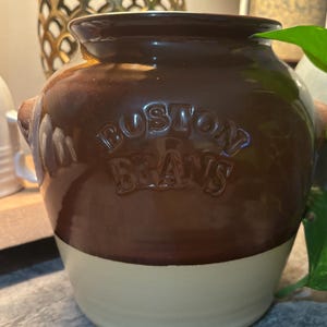May include: A brown and cream-colored ceramic Boston Beans pot with a rounded shape and two small handles. The words "BOSTON BEANS" are embossed on the front. The pot has a glossy finish.