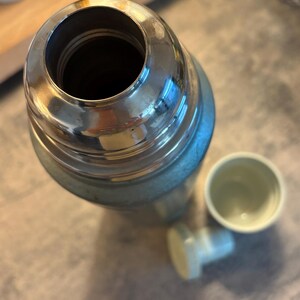 K&ouml;nnte beinhalten: Eine Vintage-Thermoskanne mit einem silberfarbenen Metalloberteil und einem hellblauen K&ouml;rper. Die Thermoskanne ist ge&ouml;ffnet und zeigt das Innere. Ein kleiner, hellfarbiger Becher und ein Verschluss sind neben der Thermoskanne.