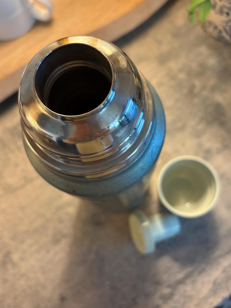K&ouml;nnte beinhalten: Eine Vintage-Thermoskanne mit einem silberfarbenen Metalloberteil und einem hellblauen K&ouml;rper. Die Thermoskanne ist ge&ouml;ffnet und zeigt das Innere. Ein kleiner, hellfarbiger Becher und ein Verschluss sind neben der Thermoskanne.