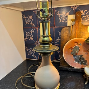 May include: A vintage table lamp with a beige, bulbous base and brass accents. The lamp features a round, frosted glass lightbulb and a brass shade holder. The lamp is plugged in and lit, casting a warm glow. The lamp is approximately 18 inches tall.