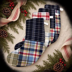 May include: A set of four quilted fabric Christmas stockings featuring various plaid patterns in navy blue, red, green, and white. Each stocking includes a small white fabric loop at the top for hanging, showcasing a patchwork design.