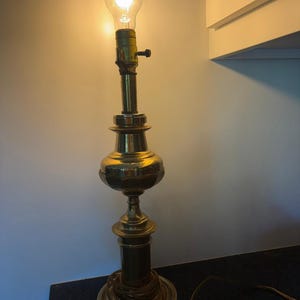 May include: A vintage brass table lamp with a lit Edison bulb. The lamp has a decorative, tiered base and a long, slender neck. The lamp is approximately 24 inches tall.