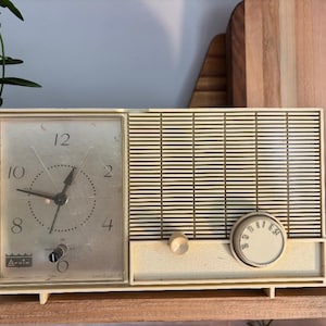 May include: Vintage off-white alarm clock with a clock face on the left and a radio-style speaker on the right. The clock face has black hands and numbers. The clock is labeled "Arvin".