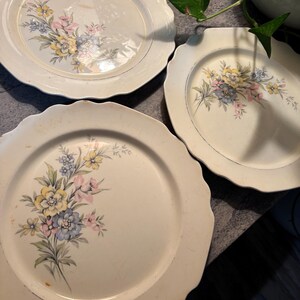 May include: Three off-white ceramic plates with scalloped edges. Each plate features a floral design in yellow, pink, and blue. The plates are arranged on a dark surface, with a plant visible in the background.