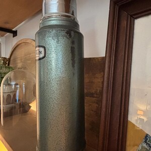 K&ouml;nnte beinhalten: Vintage Stanley Thermoskanne in gesprenkeltem Gr&uuml;n mit gl&auml;nzendem silbernem Deckel und Boden. Die Thermoskanne hat ein schwarz-wei&szlig;es Etikett. Ein klassischer Isolierbeh&auml;lter f&uuml;r hei&szlig;e oder kalte Getr&auml;nke.