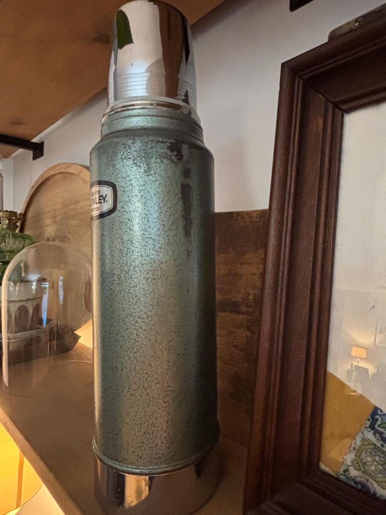 K&ouml;nnte beinhalten: Vintage Stanley Thermoskanne in gesprenkeltem Gr&uuml;n mit gl&auml;nzendem silbernem Deckel und Boden. Die Thermoskanne hat ein schwarz-wei&szlig;es Etikett. Ein klassischer Isolierbeh&auml;lter f&uuml;r hei&szlig;e oder kalte Getr&auml;nke.