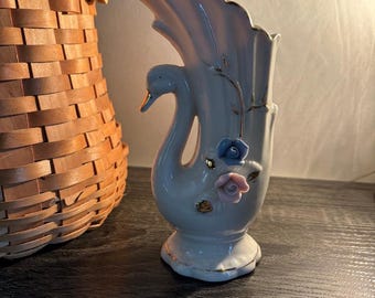 May include: A white ceramic swan vase with a gold trim and accents. The vase features a swan neck and a fan-shaped back. It is decorated with blue and pink flowers.