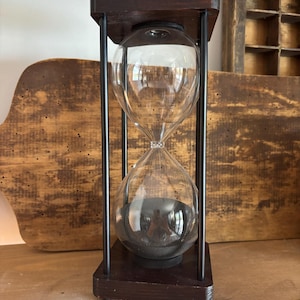 May include: An hourglass with a dark wood frame and black metal supports. The clear glass bulbs contain dark sand. The hourglass is a decorative item, suitable for home decor or as a functional timer.