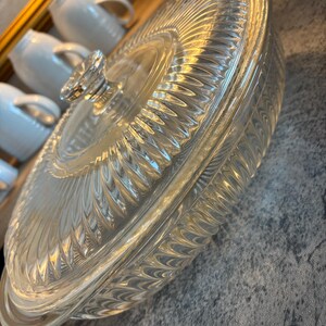 May include: Clear glass oval casserole dish with a lid. The dish has a textured, ribbed design and a clear glass knob on the lid. The dish is likely vintage and suitable for serving or baking.