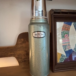 K&ouml;nnte beinhalten: Eine Vintage-Stanley-Thermoskanne in gesprenkeltem Gr&uuml;n mit gl&auml;nzendem silbernen Deckel und Boden. Die Thermoskanne hat ein schwarz-wei&szlig;es Etikett mit der Aufschrift "Aladdin's Stanley thermos". Die Thermoskanne steht auf einem Holzregal.
