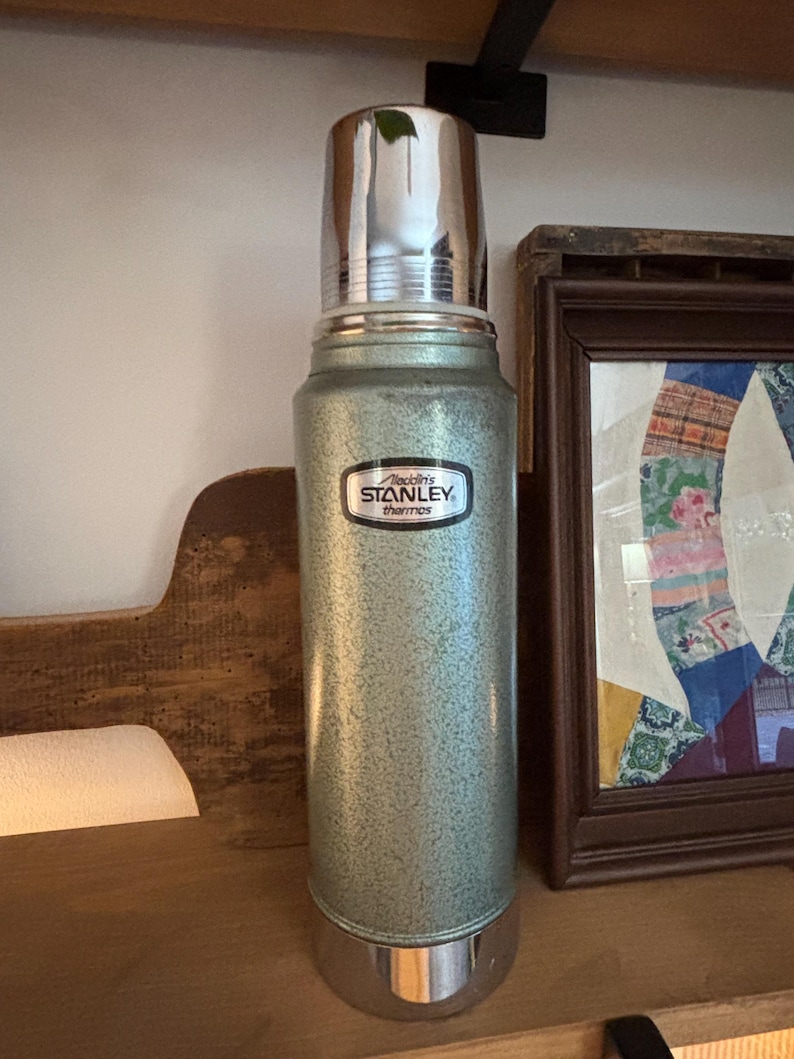 K&ouml;nnte beinhalten: Eine Vintage-Stanley-Thermoskanne in gesprenkeltem Gr&uuml;n mit gl&auml;nzendem silbernen Deckel und Boden. Die Thermoskanne hat ein schwarz-wei&szlig;es Etikett mit der Aufschrift "Aladdin's Stanley thermos". Die Thermoskanne steht auf einem Holzregal.