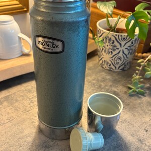 K&ouml;nnte beinhalten: Eine Vintage-Stanley-Thermoskanne in Teal mit silbernem Deckel und Boden sowie einer passenden Tasse. Auf der Thermoskanne steht "Aladdin's Stanley thermos". Der Deckel ist abgenommen, und die Tasse steht daneben.