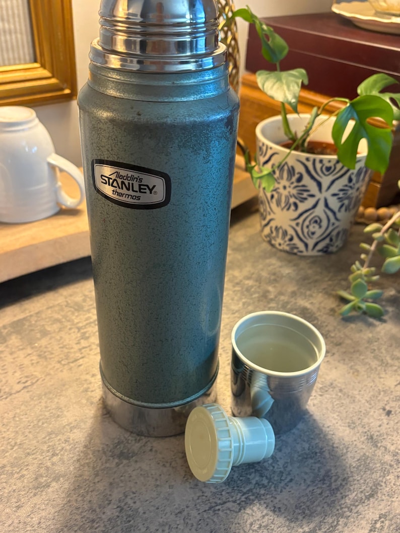 K&ouml;nnte beinhalten: Eine Vintage-Stanley-Thermoskanne in Teal mit silbernem Deckel und Boden sowie einer passenden Tasse. Auf der Thermoskanne steht "Aladdin's Stanley thermos". Der Deckel ist abgenommen, und die Tasse steht daneben.
