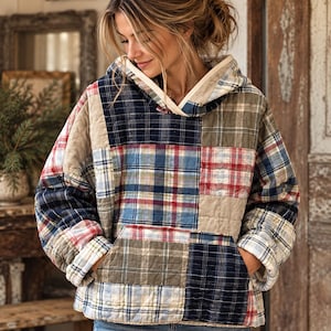 Op de afbeelding: Een gewatteerde hoodie met een patchworkontwerp van diverse ruitpatronen in tinten marineblauw, rood, wit en beige. Het kledingstuk heeft lange mouwen, een capuchon met een lichtgekleurde rand en een grote kangoeroezak aan de voorzijde.
