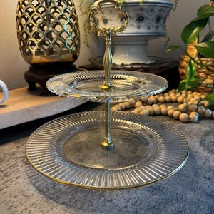 May include: A two-tiered serving tray with clear glass plates and a gold-colored metal handle. The plates have a ribbed design and gold rims. The handle has an ornate design. The tray is suitable for serving desserts or appetizers.