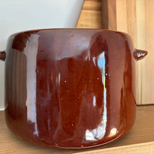 May include: A brown ceramic pot with a glossy finish. The pot has a cylindrical shape with two small handles on either side. It sits on a wooden shelf, with a wooden cutting board in the background.