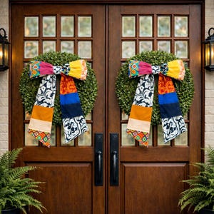 May include: Two green wreaths with colorful fabric bows hang on a wooden door. The bows feature a patchwork design with vibrant colors like pink, yellow, orange, blue, and black and white patterns. The door has glass panels.