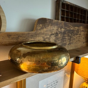 May include: A hammered brass bowl with a wide, rounded shape. The bowl sits on a wooden shelf, reflecting light. The surface has a textured, dimpled appearance. The background includes a wooden cutting board and a framed picture.