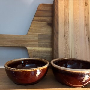 May include: Two brown ceramic bowls with a glossy finish and a light brown rim. A wooden cutting board and a wooden serving board are in the background. The bowls are on a wooden surface.