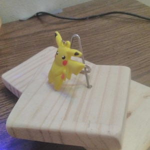 May include: A yellow Pikachu figurine with red cheeks and black eyes stands on a small, light-colored wooden block. A silver metal wire is attached to the block. The block is resting on top of another similar wooden block.
