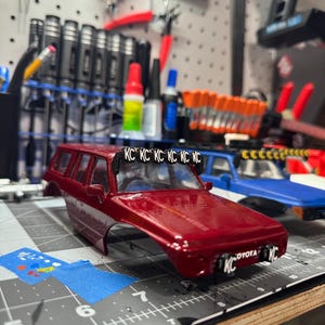 以下が含まれることがあります： 屋根にライトバー、前面に「KC」の文字が入った赤いおもちゃの車。トヨタのモデルカーです。車はカッティングマットの上にあります。