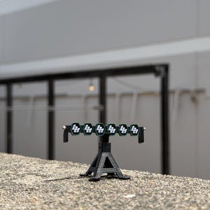 Peut inclure: Une barre lumineuse noire à six lumières avec un support. Chaque lumière a un logo blanc sur un hexagone vert. Le support est une conception noire à trois pieds. L'objet est sur une surface grise texturée.
