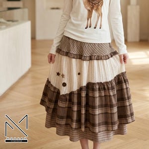 May include: A long, tiered skirt featuring alternating panels of brown plaid fabric and cream-colored knit fabric. The skirt includes decorative brown buttons and star-shaped accents on the knit section, with ruffled edges between the layers.