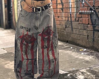 May include: Gray baggy jeans with red and black graphic designs. The jeans feature a wide leg and a black belt with silver studs. The word "HATARI" is printed on the front of the jeans.