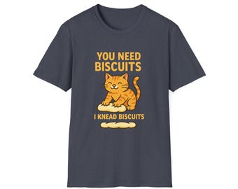 May include: A navy blue t-shirt features a cartoon cat kneading dough with the text "YOU NEED BISCUITS" and "I KNEAD BISCUITS" in yellow. The cat is orange with brown stripes and a smiling face.
