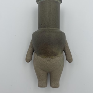 May include: A ceramic figurine shaped like a person, featuring a cylindrical head and torso, small arms, and legs. The head and upper torso are dark green, contrasting with the lighter tan lower body. The figurine has a matte finish.