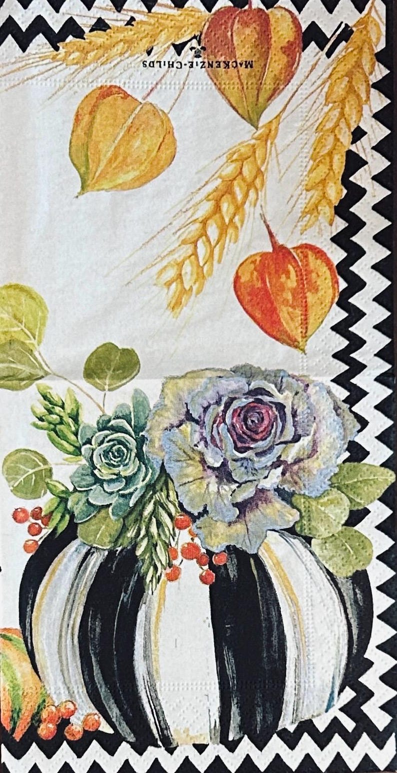 May include: A paper napkin featuring a black and white chevron border and a central illustration of a black and white striped pumpkin topped with a succulent, cabbage, red berries, wheat stalks, and orange physalis with MacKenzie-Childs text.