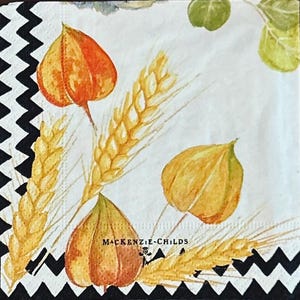 May include: A square paper napkin featuring a white background with a black zigzag border. The design includes watercolor illustrations of golden wheat stalks, orange and yellow physalis husks, and green leaves. The text MACKENZIE-CHILDS is printed.