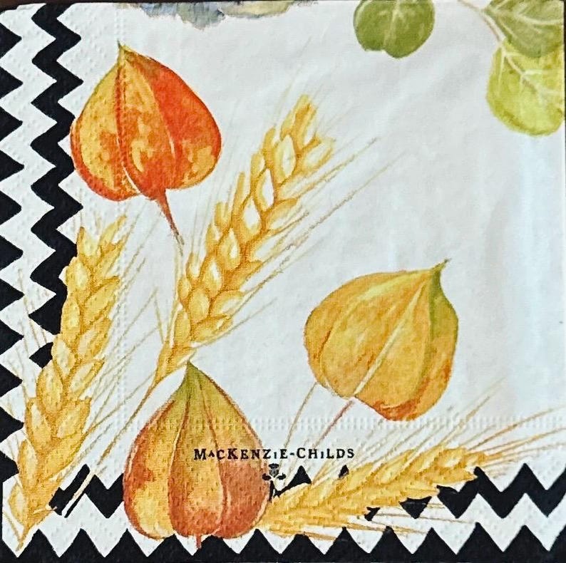 May include: A square paper napkin featuring a white background with a black zigzag border. The design includes watercolor illustrations of golden wheat stalks, orange and yellow physalis husks, and green leaves. The text MACKENZIE-CHILDS is printed.