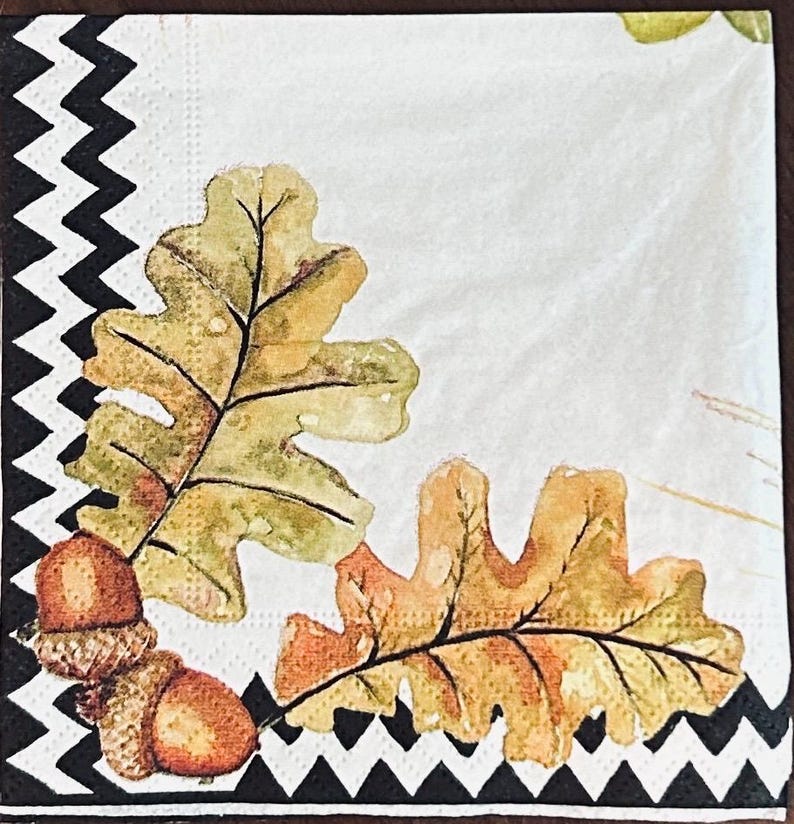May include: A square paper napkin featuring a white background with a black and white chevron border along the left and bottom edges. The design includes two watercolor-style oak leaves in shades of green and gold, alongside two brown acorns.