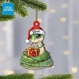 Pode incluir: Enfeite de Natal de uma cobra verde com um gorro de Papai Noel com o nome "Steve". A cobra está enrolada em volta de um presente e decorada com luzes coloridas. O enfeite está pendurado em uma corrente dourada.